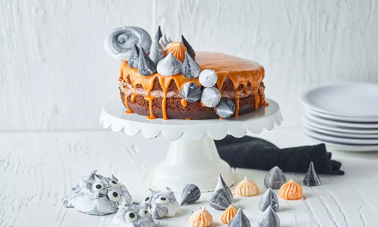 Tummanruskea halloween-kakku, oranssilla valuvalla kuorrutteella, mustia ja harmaita marenkeja koristeena valkoisella jalustalla