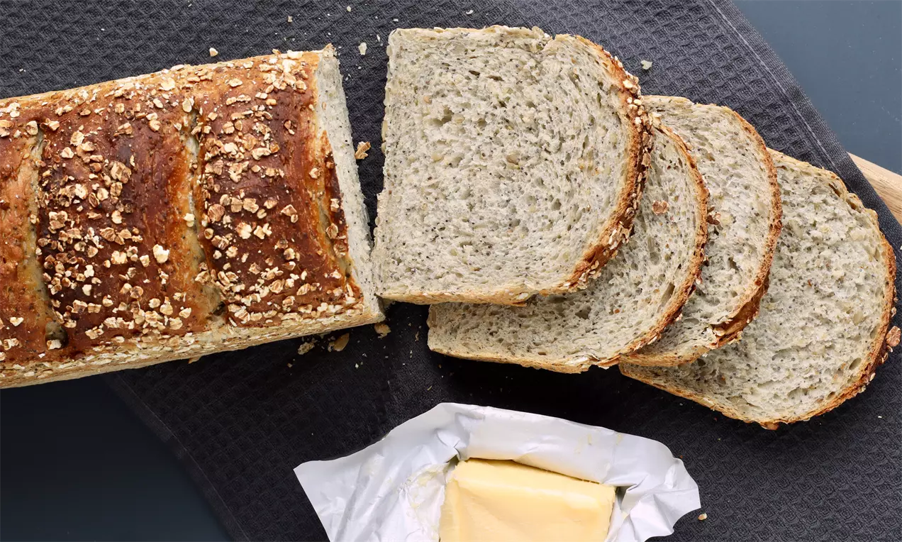 Vaaleanruskean kuoren ja kaurahiutaleiden peittämä vuokaleipä viipaleina tummaa kangasta vasten, vieressä avattu voi kääreessä