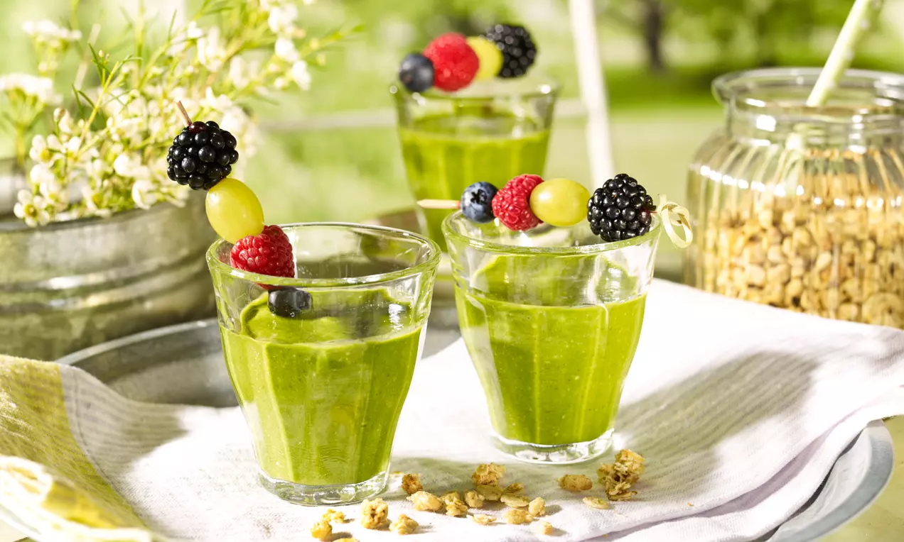 Lasissa vihreän samettista smoothieta, pinnalla marjoja vartaassa, ympärillä kultaista granolaa ja kukkia vaalealla tarjottimella