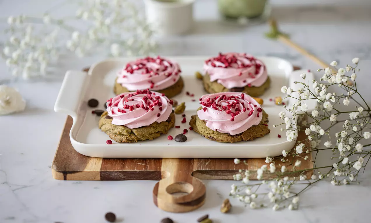 Vihreään sävyyn taittuvat pehmeät cookiet, joiden yllä vaalea vaaleanpunainen marenkivoi pursotettuna ja joukossa vadelmanpunaisia murusia