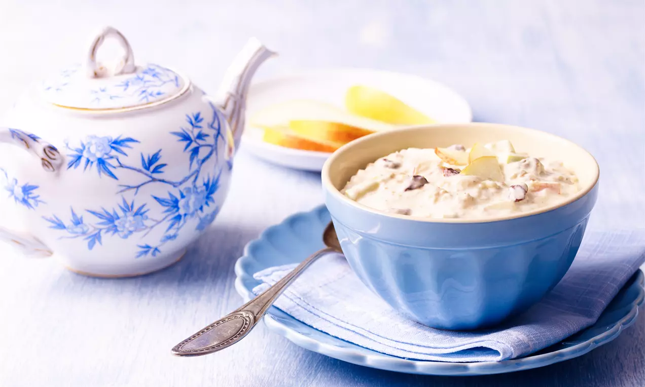 Kermanvalkoista, ruskeita ja kellertäviä pilkkuja sisältävä Bircher-mysli vaaleansinisessä kulhossa kankaan ja lautasen päällä lusikan vieressä