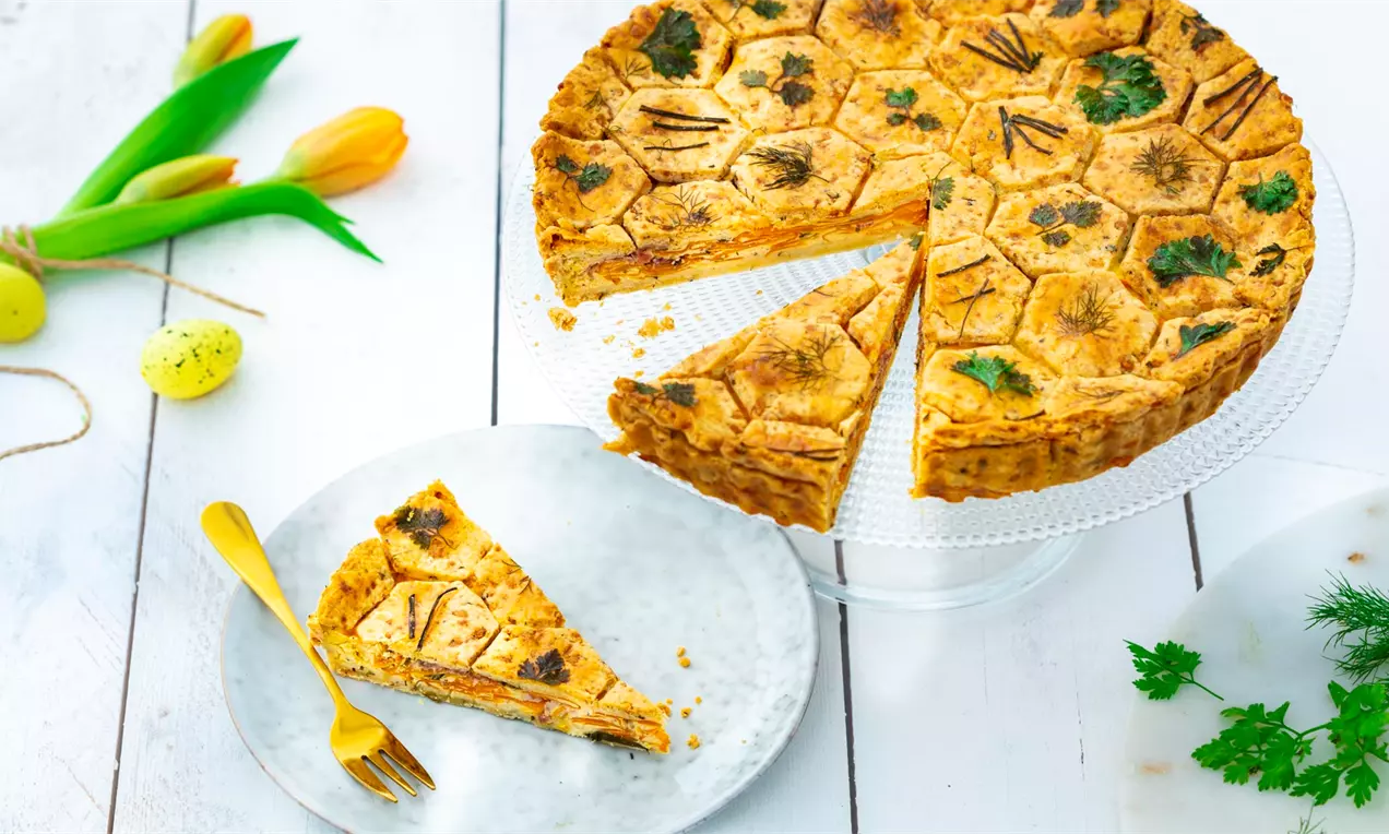 Porkkana-yrttipiirakka kullanhohtoinen ja täyteläinen, kuusikulmaiseen kuviopintaan upotettuja yrttisilmuja, lautastarjottimella ja viipaleena valkoisella lautasella