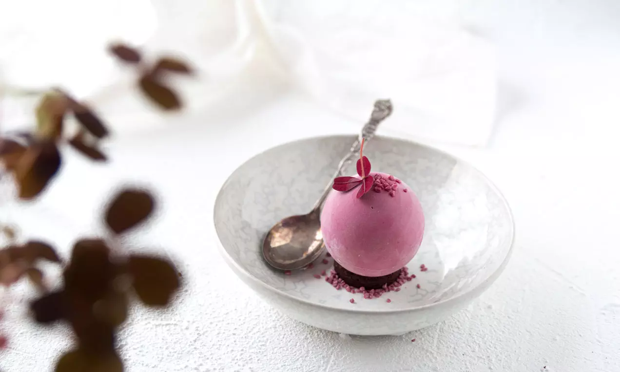 Pyöreä, sileä ja syvän violettipinkki kuula aseteltuna valkoiselle lautaselle viereen tumma lusikka ja koristeena hennot lehdet