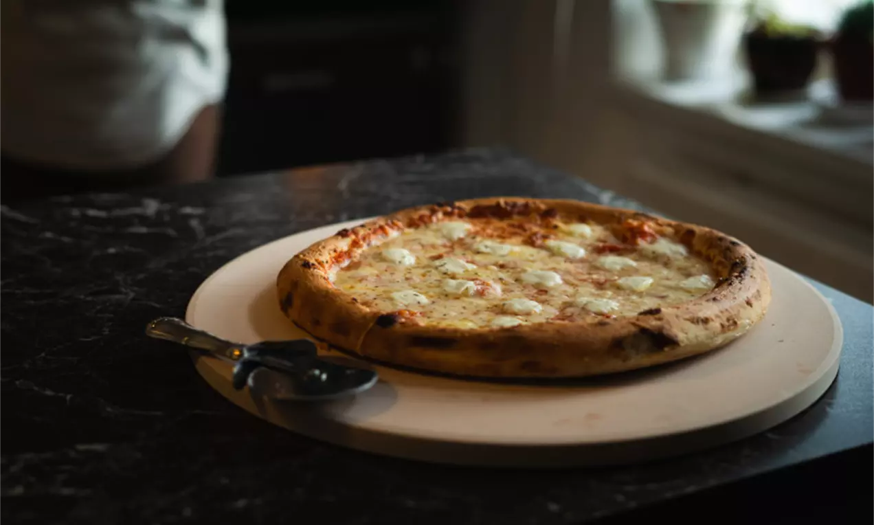 Pakastepizza kullanruskealla reunalla, sulanut juusto täytteenä, aseteltuna vaalealle pyöreälle alustalle keittiön hämärässä valossa