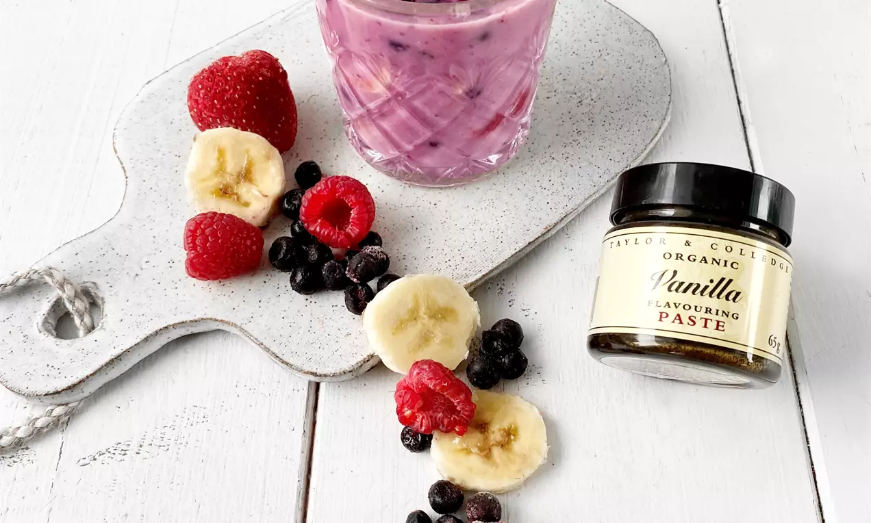 Vaaleanpunainen smoothie lasissa, pinnalla vadelma, banaaniviipale ja mintunlehti, vierellä leikkuulaudalla marjoja ja banaaniviipaleita