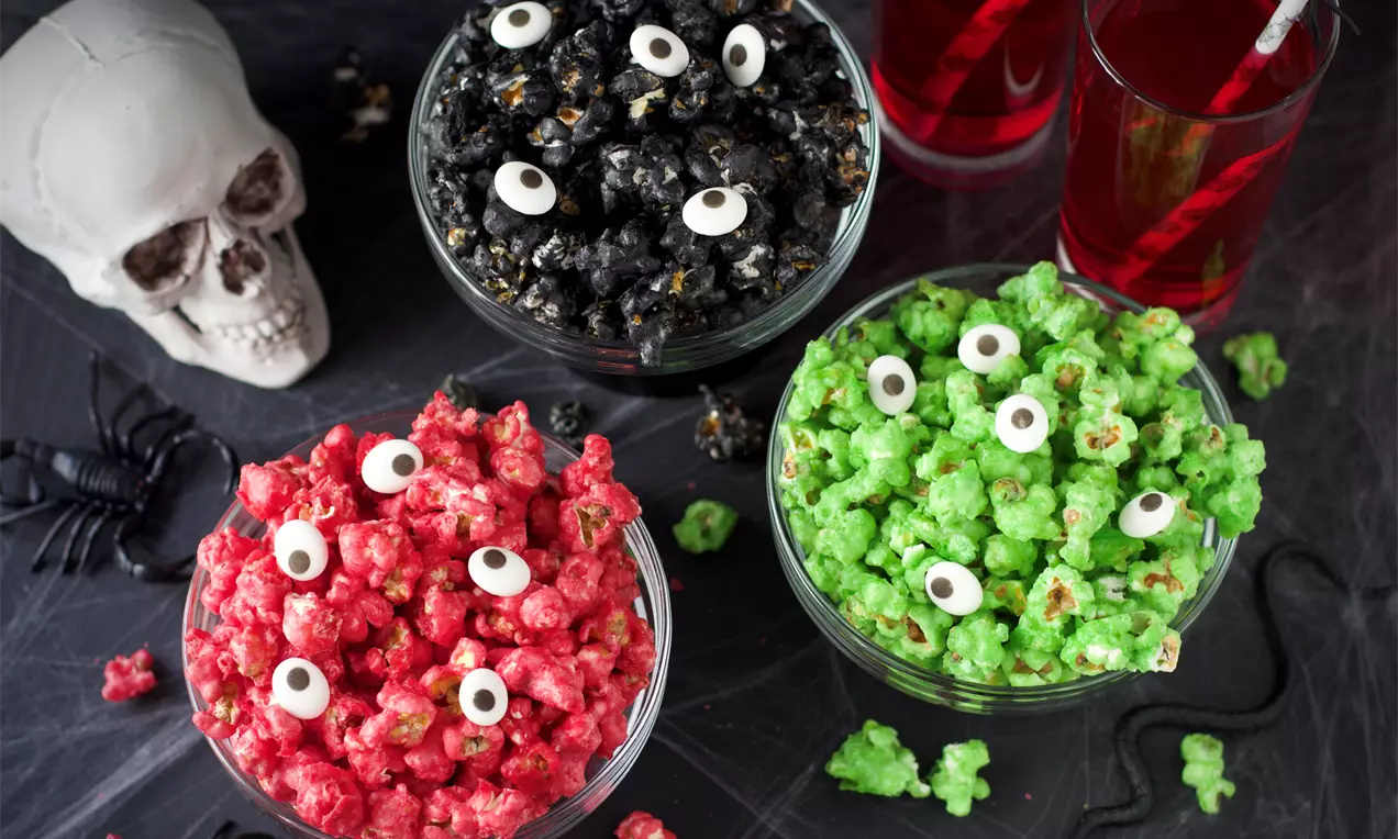 Punaiset, mustat ja vihreät popcornit lasikulhoissa, karkkisilmillä koristeltuina, aseteltuina hämähäkinverkkoiselle pöydälle halloween-rekvisiitan keskellä