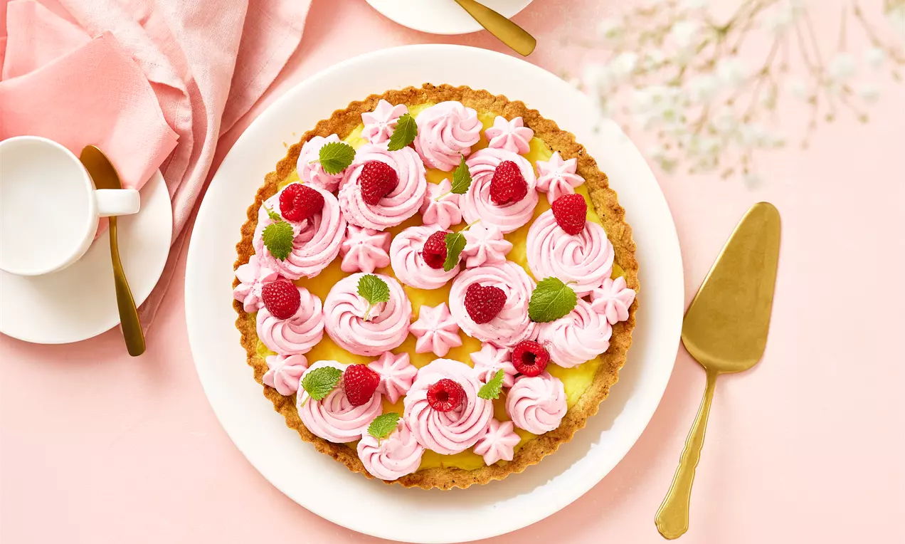 Vaaleankeltainen sitruunapiiras rapealla reunahelmalla, vaaleanpunaisen kreemin kiertyneitä pyörteitä, vadelmia ja mintunlehtiä valkoisella lautasella