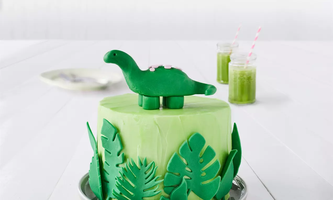 Vaaleanvihreällä kuorrutettu pyöreä kakku, koristeltu tummanvihreillä lehtikuvioilla ja kruununa vihreä dinosaurus keskellä