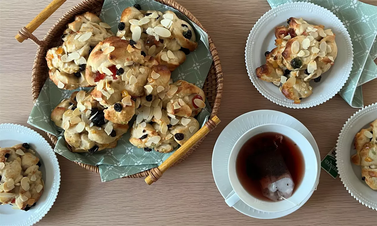 Kløbenboller-vaaleita, pehmeän kuohkeita, rusinan ja mantelilastujen täplittämiä, vihreällä kuviokankaalla korissa ja valkoisilla lautasilla teekuppien vieressä