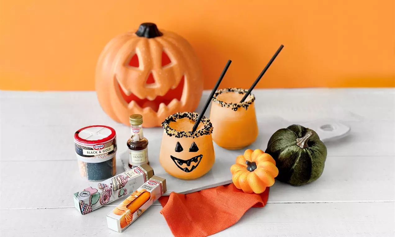 Kirkkaanoranssi pirtelö lasissa mustilla nonparellireunoilla, jack-o’-lantern-koristeet, sulkaat ja kurpitsa-asetelma valkoisella pöydällä