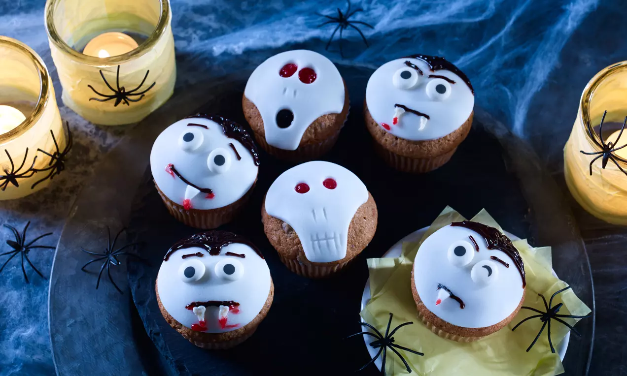 Valkoisella kuorrutteella koristellut halloweenmuffinit, vampyyri- ja pääkallokasvojen punaiset yksityiskohdat ja mustat hämähäkit tummalla tarjottimella