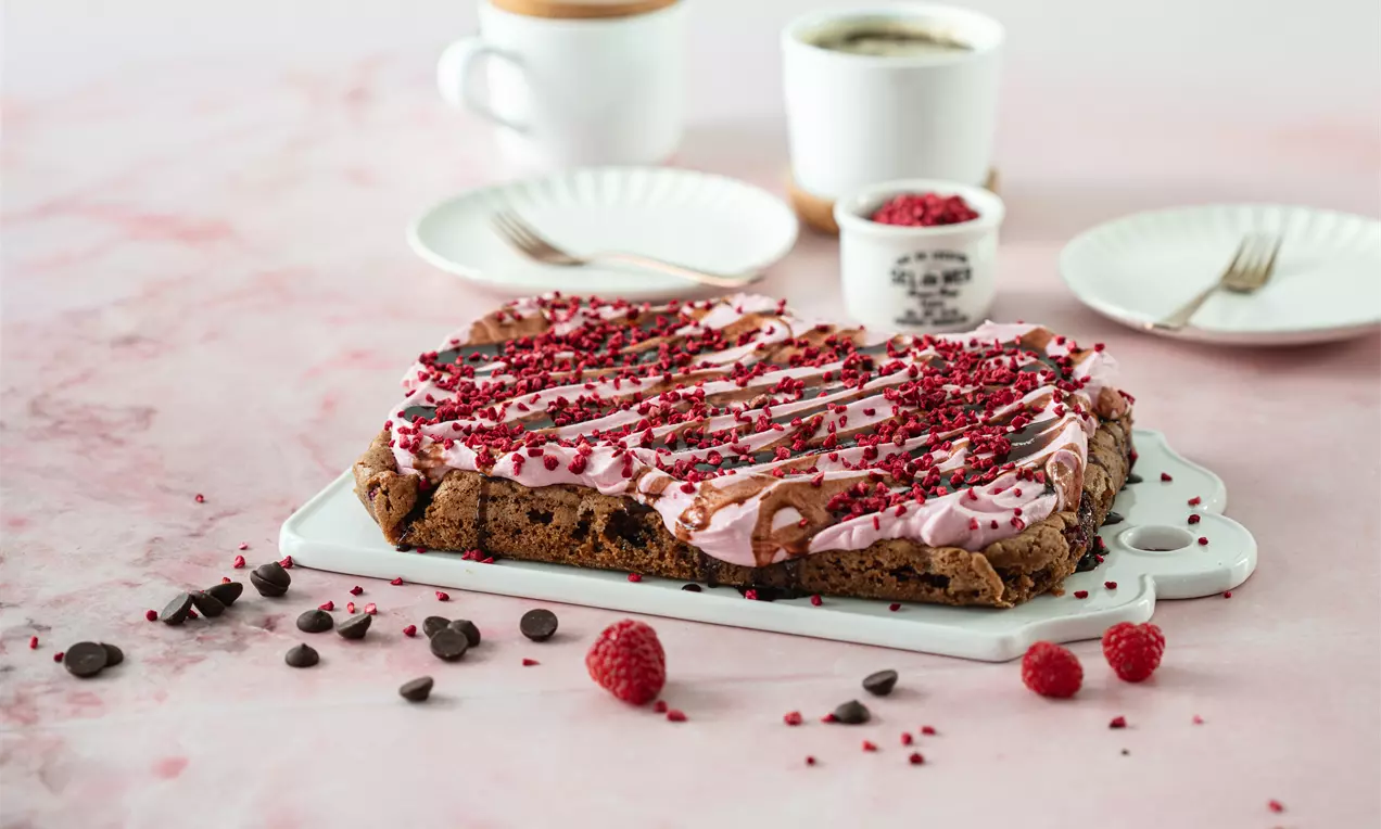 Tumma ja kostea brownie vaaleanpunaisella vadelmakuorrutteella, suklaaraidoilla ja kuivatuilla vadelmamuruilla, tarjottuna valkoisella vadilla