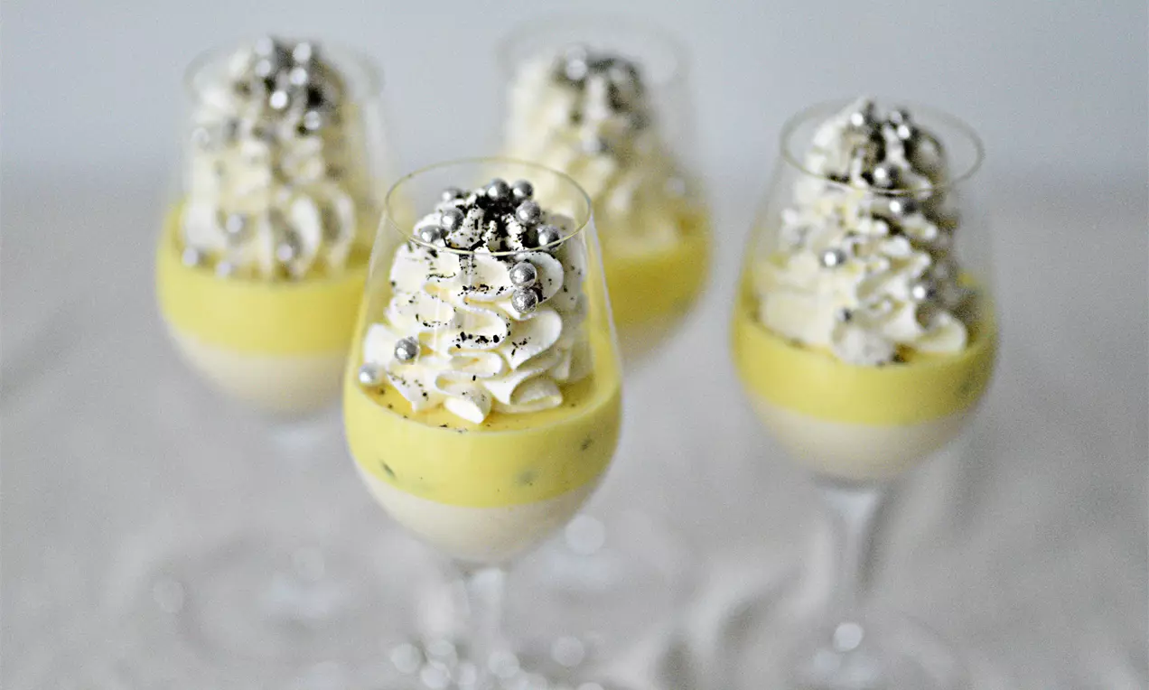 Kahden kerroksen pannacotta lasissa, vaniljanvalkoinen pohja ja kiiltävä passionhedelmän keltainen pinta, koristeltu kermavaahdolla ja hopeapalloilla
